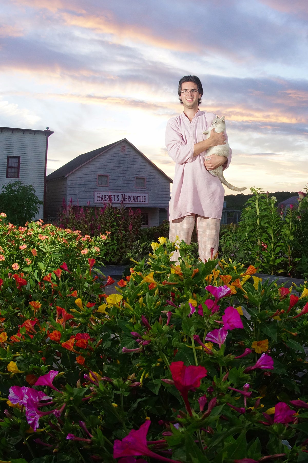 A man holding a white cat in the village flower gardens at sunset