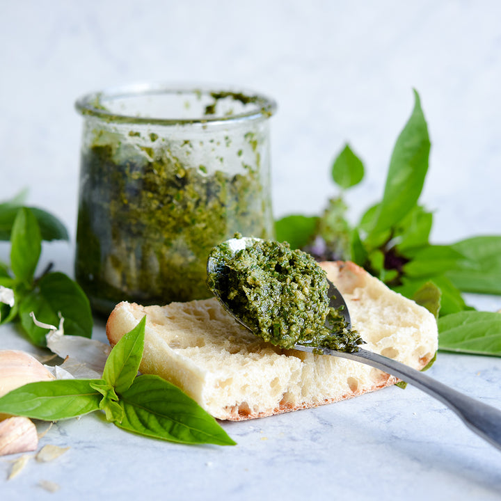 A spoonful of Siam Queen Thai Basil pesto sauce resting on a slice of artisan bread surrounded by fresh leaves.
