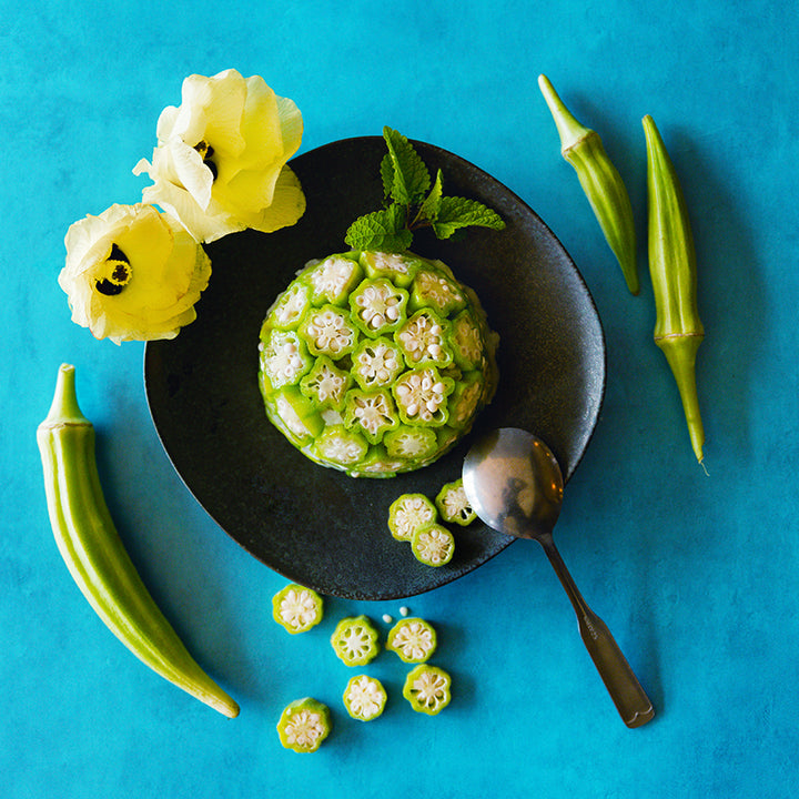 Clemson Spineless Okra slices on black plate with bloomed flowers on blue background.