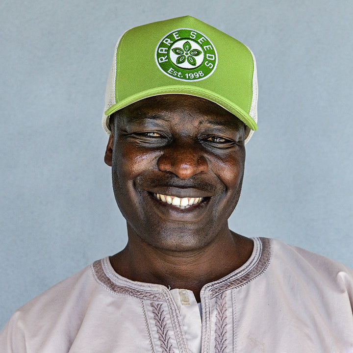 A happy smiling man wearing an Avocado Green Baker Creek Organic Cap against a grey wall.