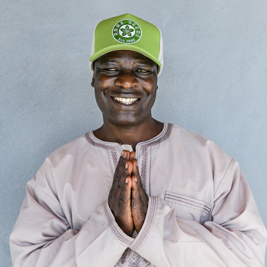 A smiling man holding his hands together while wearing an Avocado Green Baker Creek Organic Cap against a grey wall.