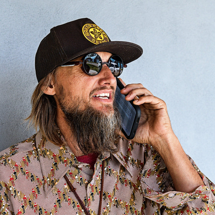 A smiling man talking on phone while wearing a Baker Creek Organic Cap against a grey wall.