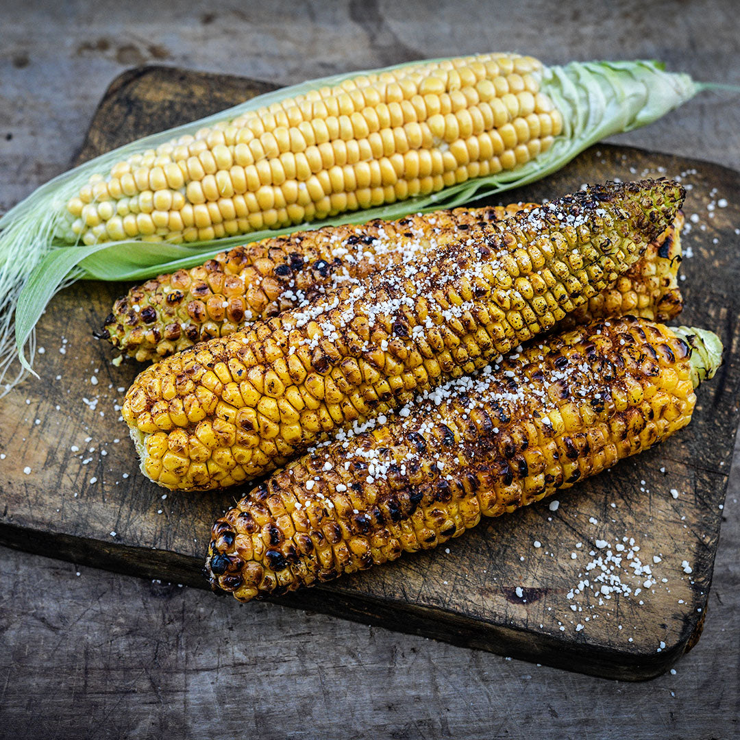 Grilled and seasoned Damaun KS Super Sweet Corn with one fresh cob on a wooden cutting board.