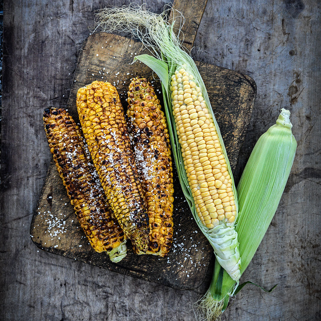 Grilled and seasoned Damaun KS Super Sweet Corn with raw cobs on a wooden background.