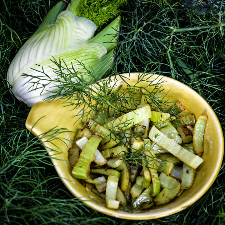 Sauteed fennel bulb in a yellow bowl with a whole bulb on a bed of fennel foliage.