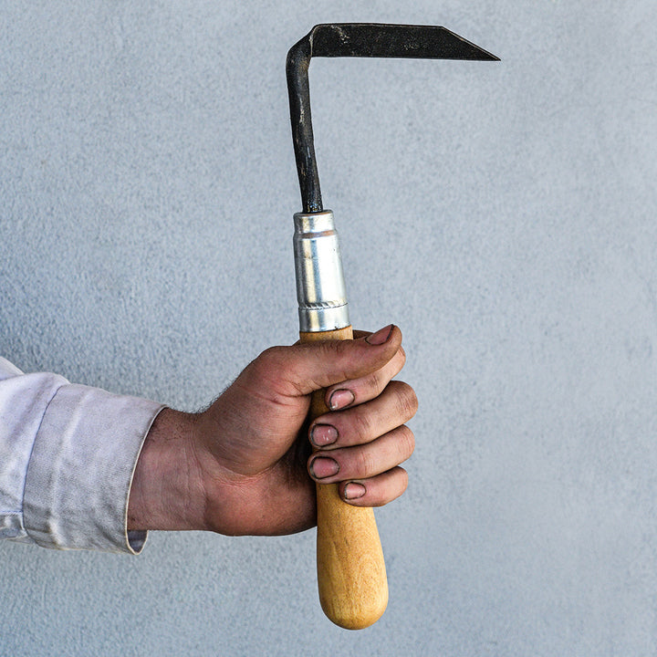 The Baker Creek blacksmith holds up a Cape Cod Left Handed Weeder against a gray wall.