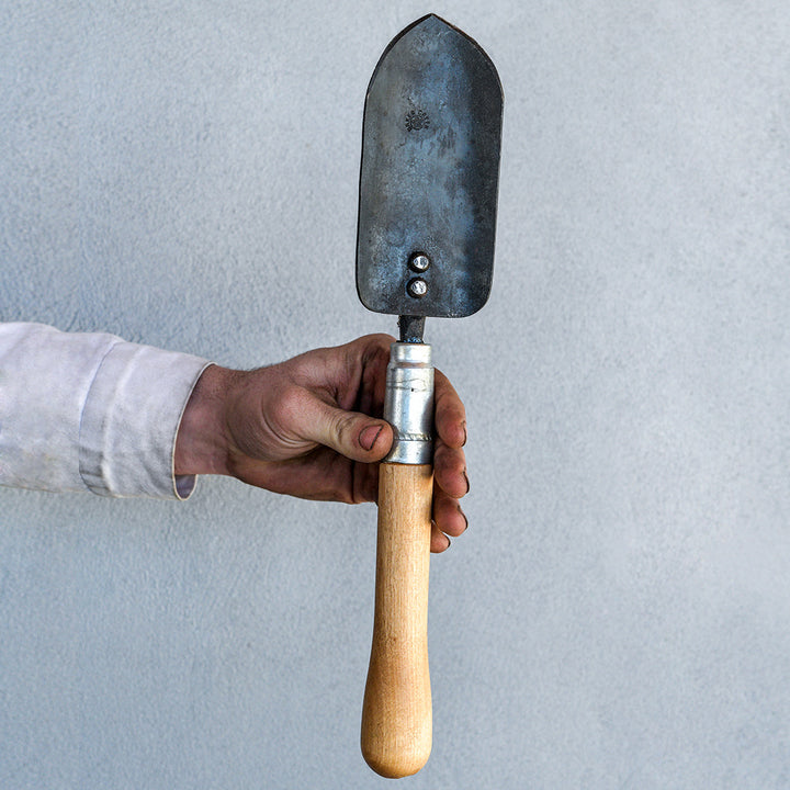 The Baker Creek blacksmith holding a Long Handled Trowel against a gray wall.