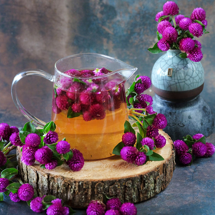 Buddy Purple Gomphrena tea in a glass teapot surrounded by blooms on a darker background.