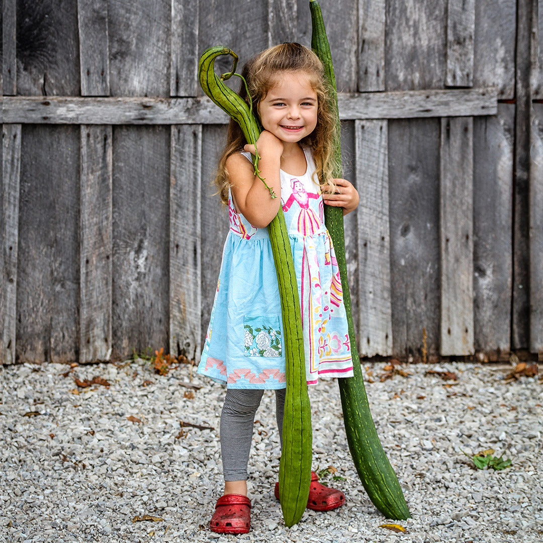 A little girl smiles big as she holds Thai Extra Long Sponge Gourds outside against a wooden wall.