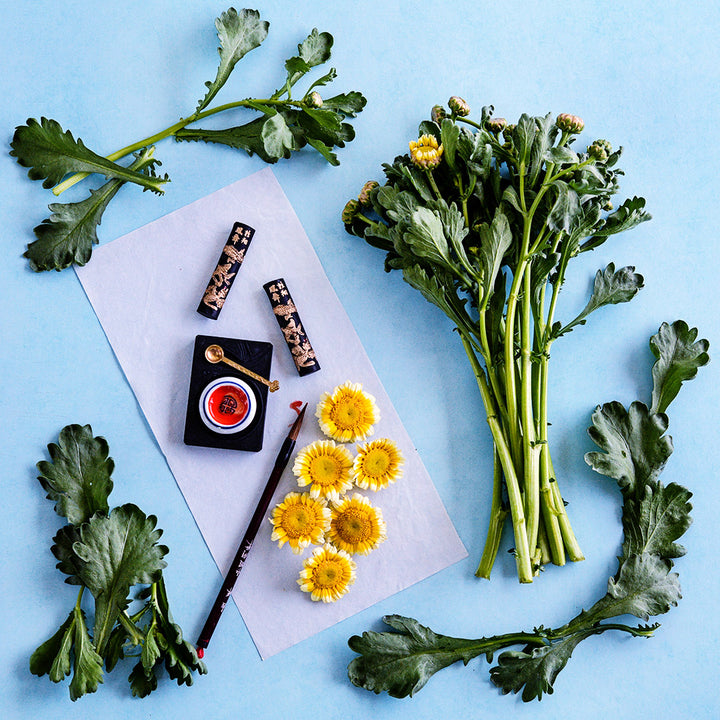 Otafuku Chrysanthemum stems and greens with flowers arranged on a light blue background.