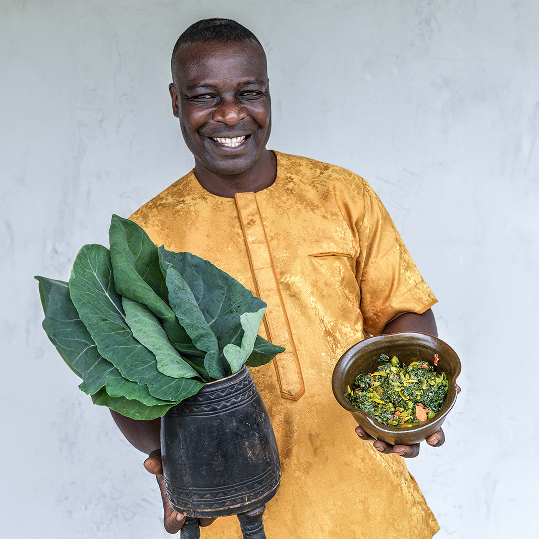 Paul from Kenya holding a vase of Sarah kale in one hand and a bowl of cooked kale in the other. Wearing a gold robe against a stone grey background.