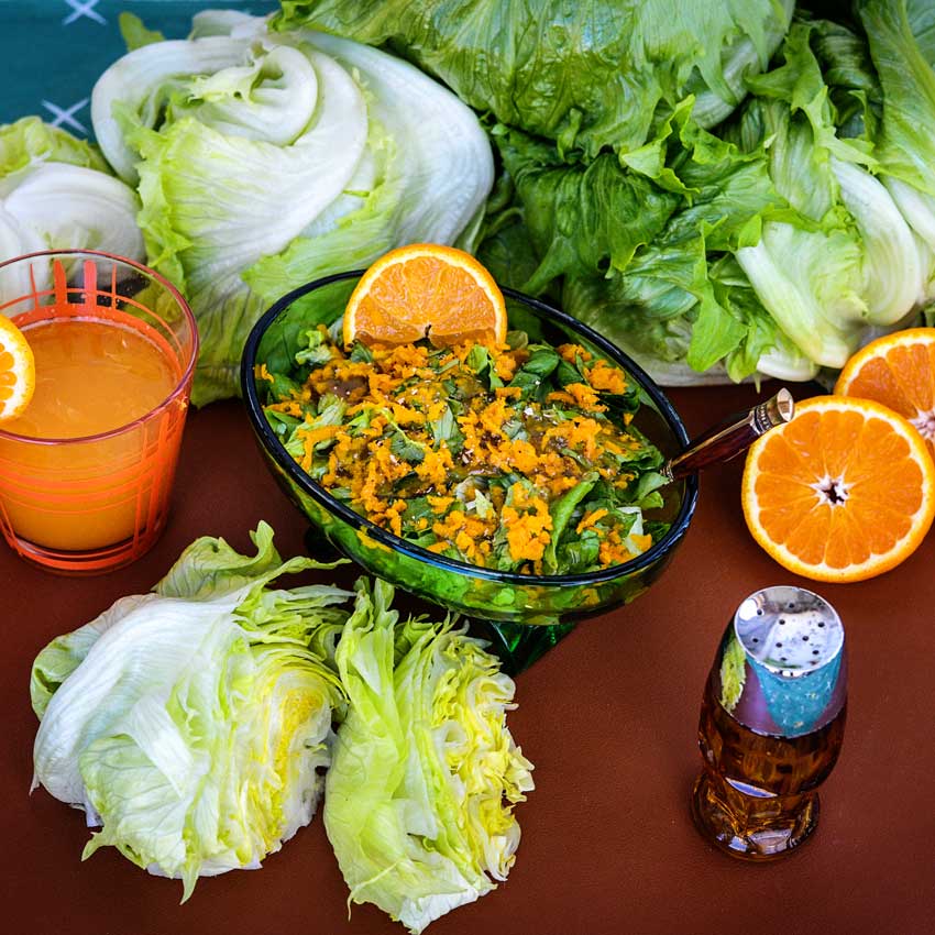 An Ice Queen lettuce salad centered on a table surrounded with sliced oranges, a cup of orange juice, and more whole heads of lettuce.