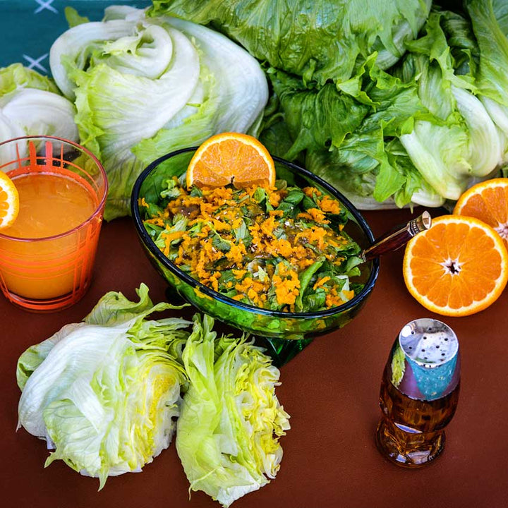 An Ice Queen lettuce salad centered on a table surrounded with sliced oranges, a cup of orange juice, and more whole heads of lettuce.