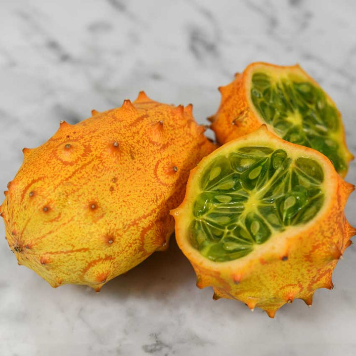 Two Kiwano African horned jelly melons, one sliced in half, the other whole on a marble background.