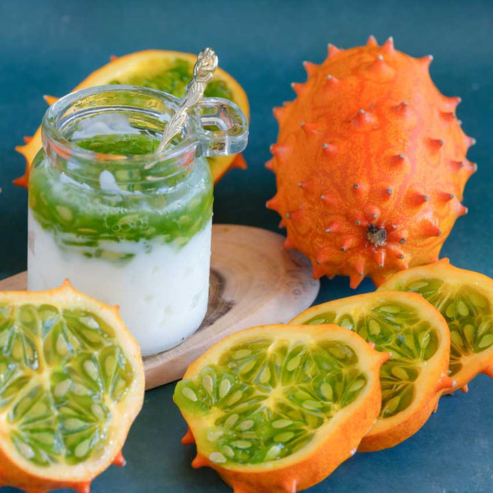Sliced and whole Kiwano African horned jelly melons next to a yogurt pudding cup topped with melon on a dark blue background.