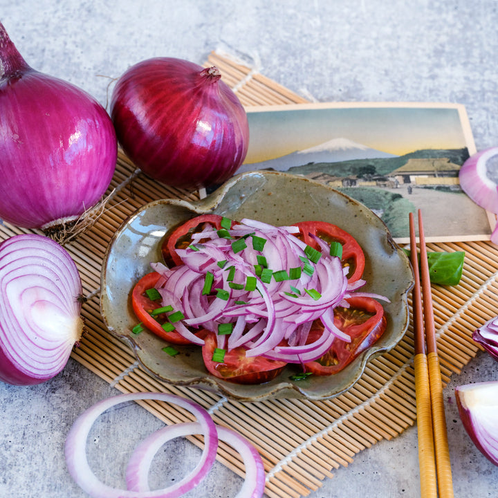 Whole and sliced Akatama Salad Red Onions with an onion tomato salad on a bamboo placemat.