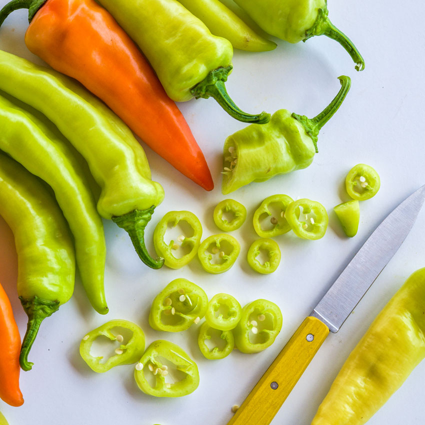 Close-up of Banana Sweet Peppers with whole and sliced pieces next to a yellow-handled peering knife on a white background.