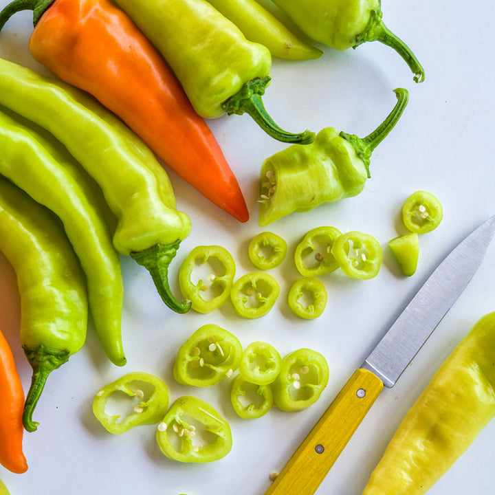 Close-up of Banana Sweet Peppers with whole and sliced pieces next to a yellow-handled peering knife on a white background.