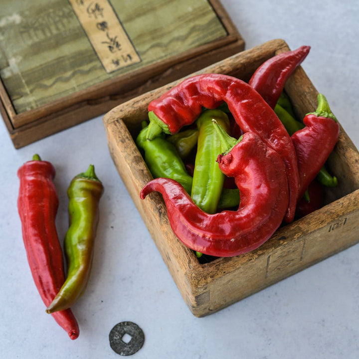 Manganji Sweet Peppers in a wooden box, others scattered on top of Japanese decor on a white background.