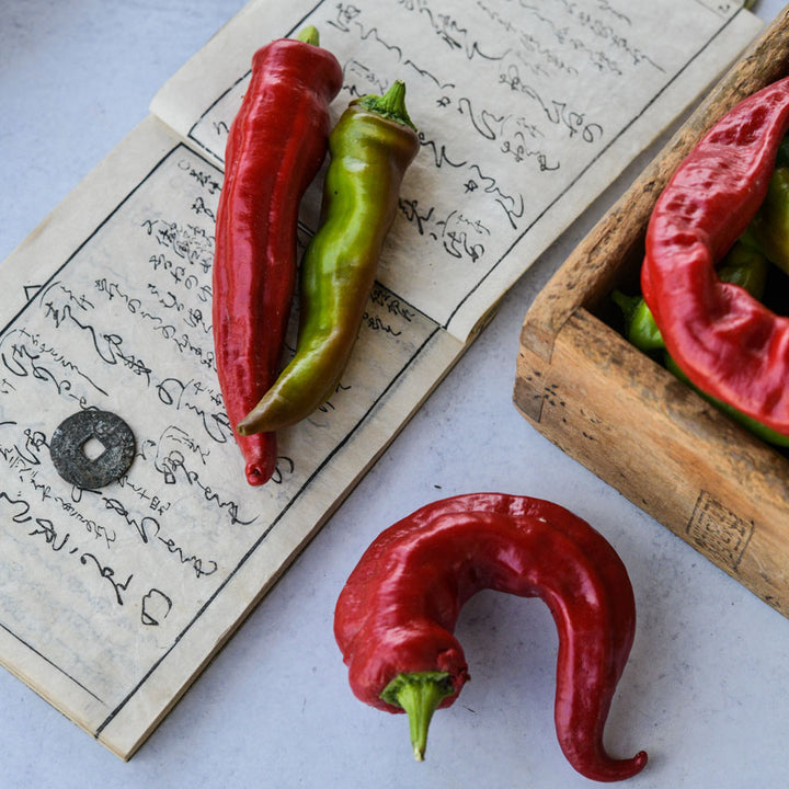 Manganji Sweet Peppers, some in a wooden box others scattered on top of Japanese decor on a white background.