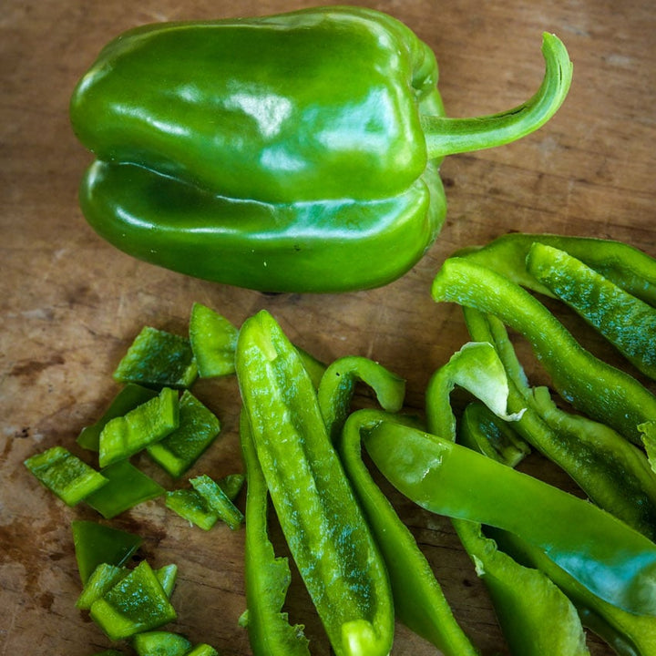 One green Ozark Giant Pepper next to sliced and chopped pepper on a wooden block.