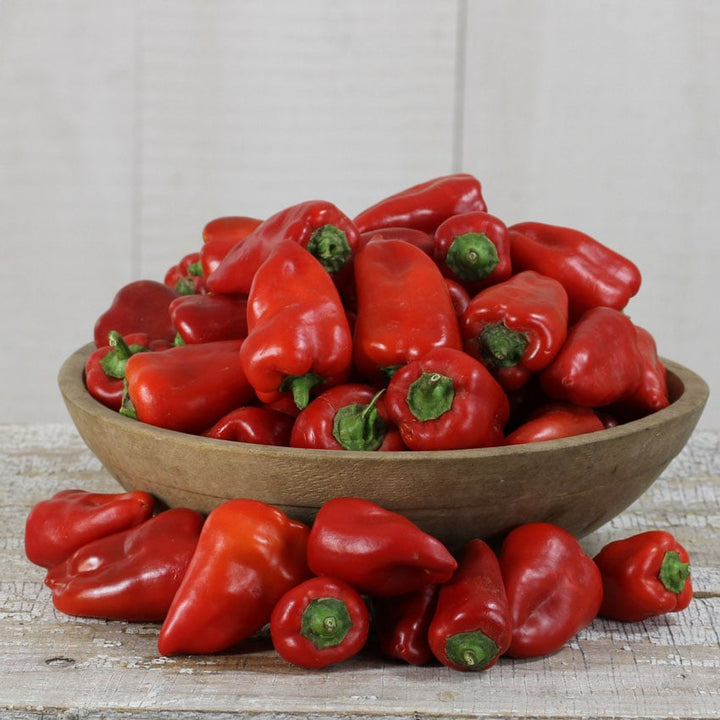 Several red Lipstick Peppers in a wooden bowl surrounded by more red peppers on a white background.