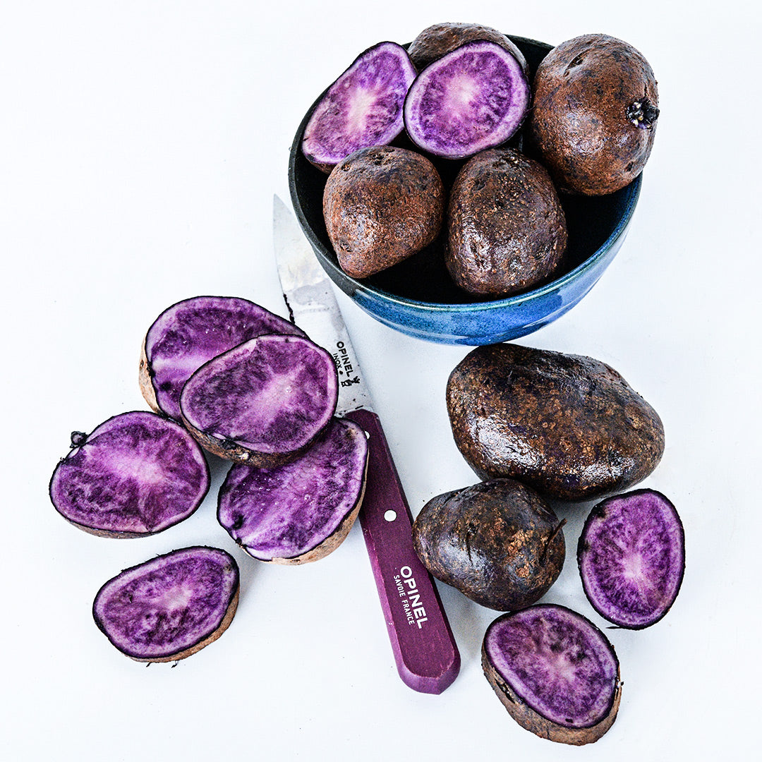 Bora Valley Mini Potatoes in a small ceramic bowl surrounded by cut and whole potatoes with a knife.