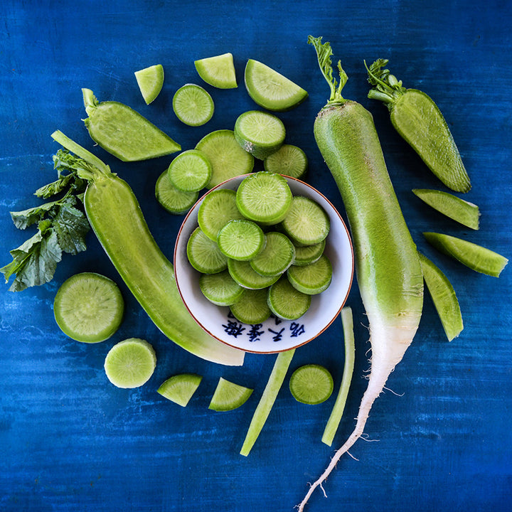Whole and cut Chinese Shawo Fruit Radishes arranged in and around a bowl on dark blue background.