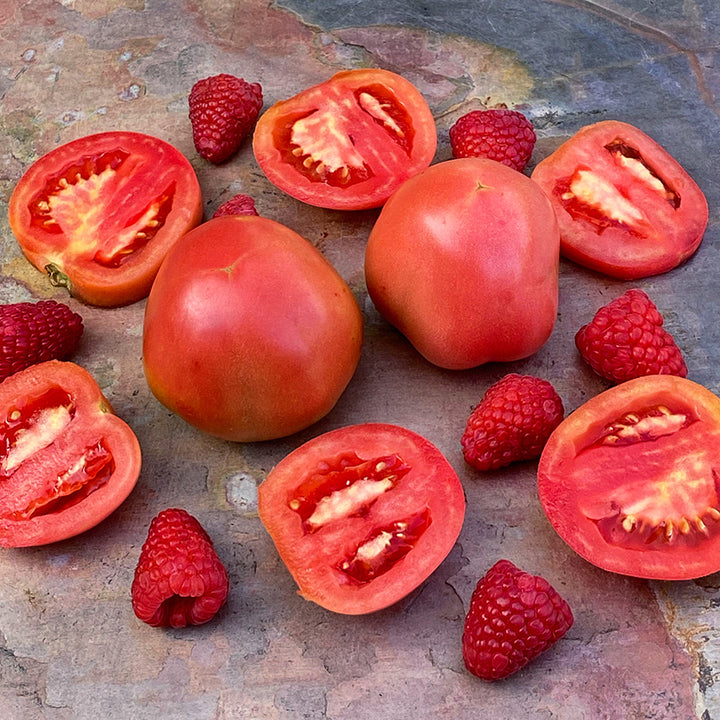Cut and whole Raspberry Lyanna Tomatoes with raspberries on a slate background.