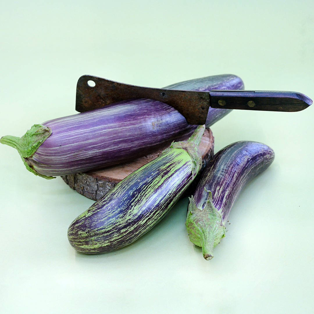 A trio of Niranjan Eggplants with a butcher knife on a light green background.
