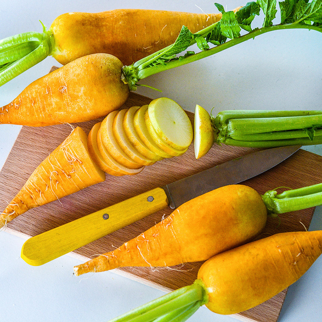 Cut and whole Lutea Carrot Radishes with a yellow handled knife on cutting board.