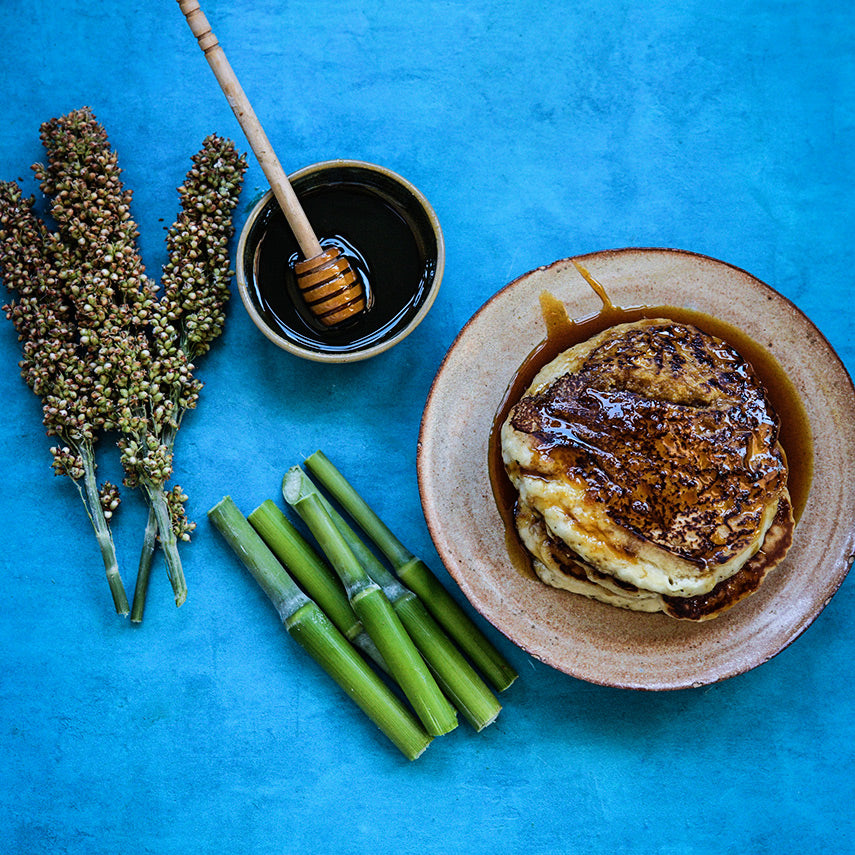 Seed heads and canes of Honey Drip Sorghum with sorghum syrup and pancakes on a bright blue background.