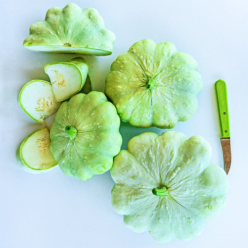 Cut and whole Bennings Green Tint Scallop Summer Squash with a green handled knife on a white background.