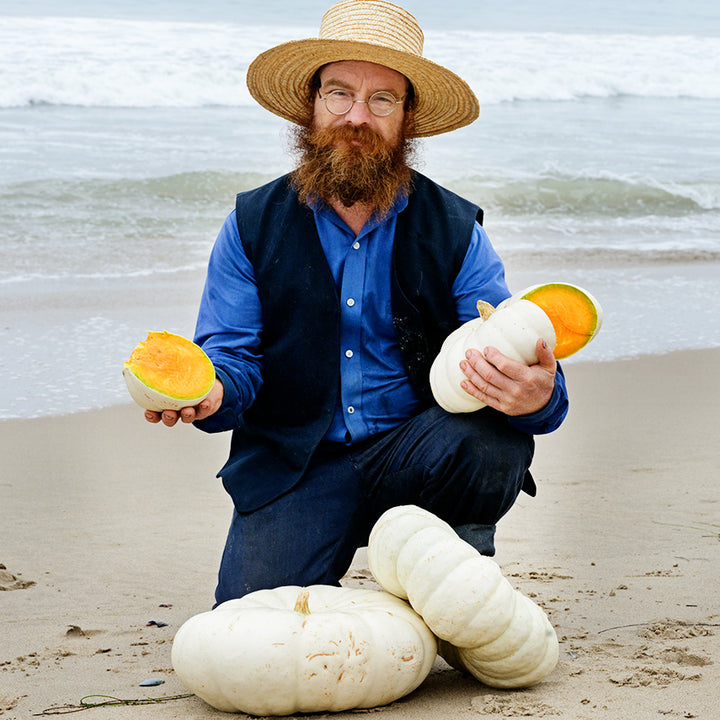 Man holding a cut Flat White Boer Pumpkin Winter Squash with a couple of pumpkins at his feet on the beach.