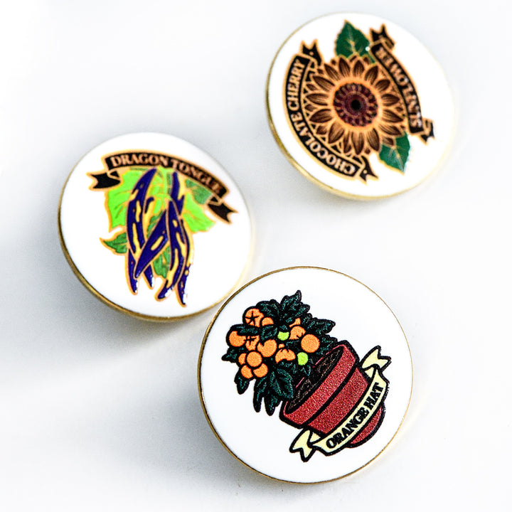 Three Limited Edition Pins with the Orange Hat Tomato Limited Edition Pin in the foreground.