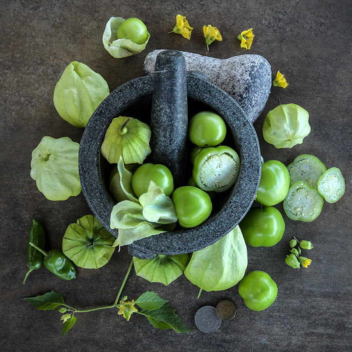 Rio Grande Verde Tomatillos in a mortar with a pestle surrounded by tomatillos, blooms and leaves.