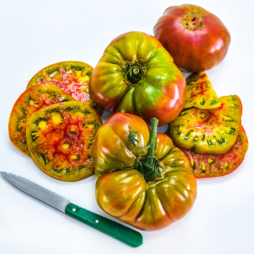 Whole and cut Ananas Noire or Black Pineapple Tomatoes with a green handled knife on a white background.