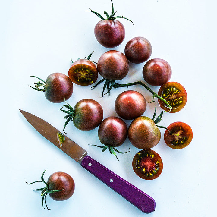 A cluster of halved and whole Black Cherry Tomatoes on a light background with a purple handled knife.