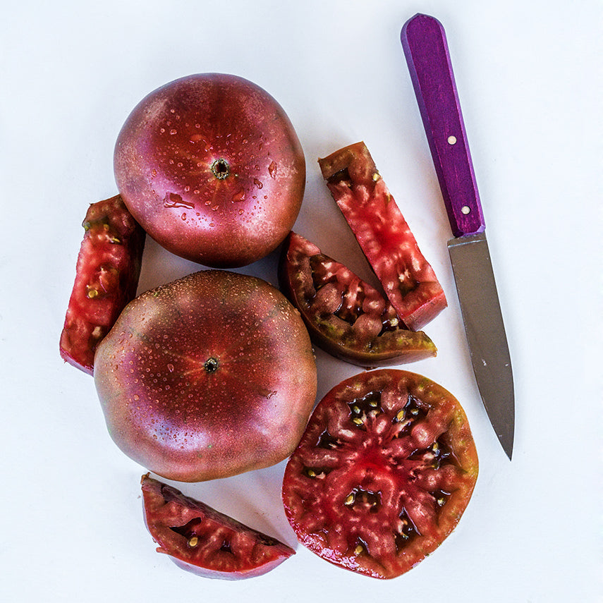 Cut and whole Black Krim Tomatoes arranged with a purple handled knife on white background.