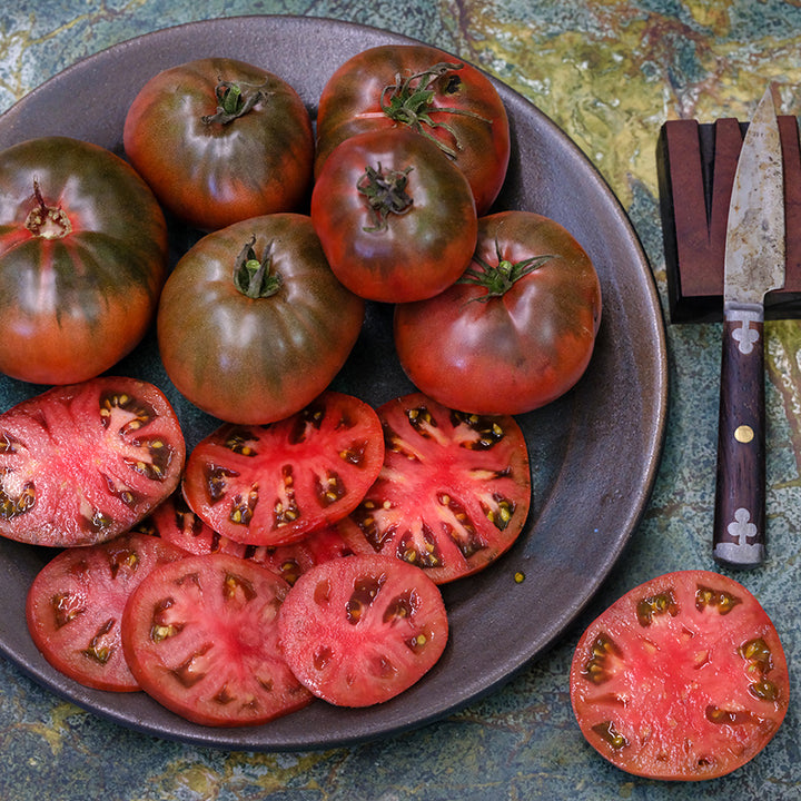 A half dozen whole Carbon Tomatoes with slices arranged on plate with a knife.
