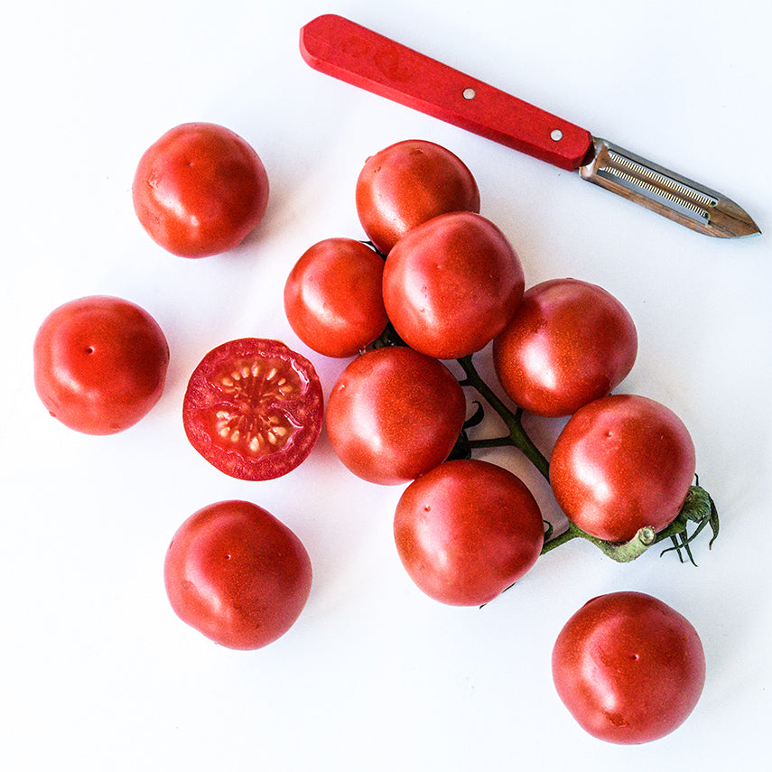 A cluster of cut and whole Chadwick Cherry or Camp Joy Tomatoes with a red handled peeler on white.