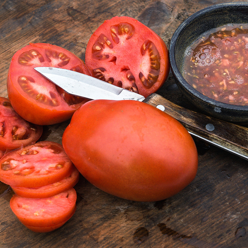 A whole Comstock Sauce and Slice Tomato with slices and juice in small bowl on a wood cutting board.