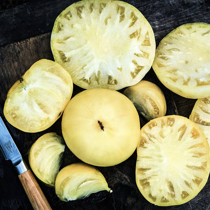 Whle and sliced Great White Tomatoes on a wooden cutting board with a knife.