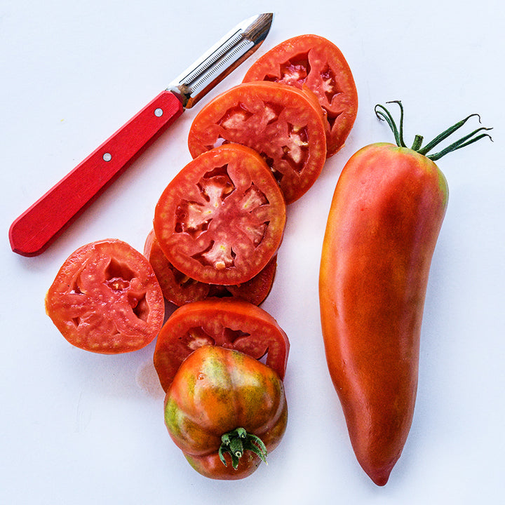 One whole and one sliced Jersey Devil tomato with a red handled peeler on a white background.