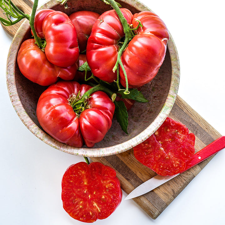 A bowl full of Mortgage Lifter Tomatoes with cut tomato on a wooden cutting board.