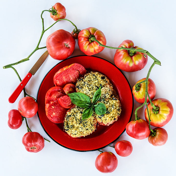 Baked Mortgage Lifter Tomatoes on bright red plate surrounded with tomatoes one a white background.