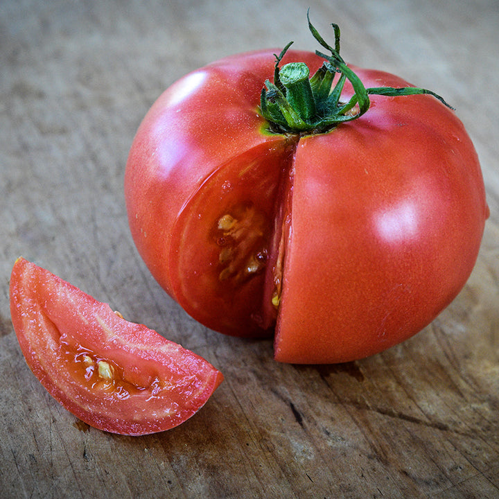 A single Ozark Pink Tomato with a slice cut on a wooden cutting board.