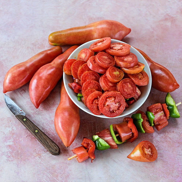 Sliced and whole Pink Fang Tomatoes in a bowl and a shish kabob with peppers and tomatoes on pink.