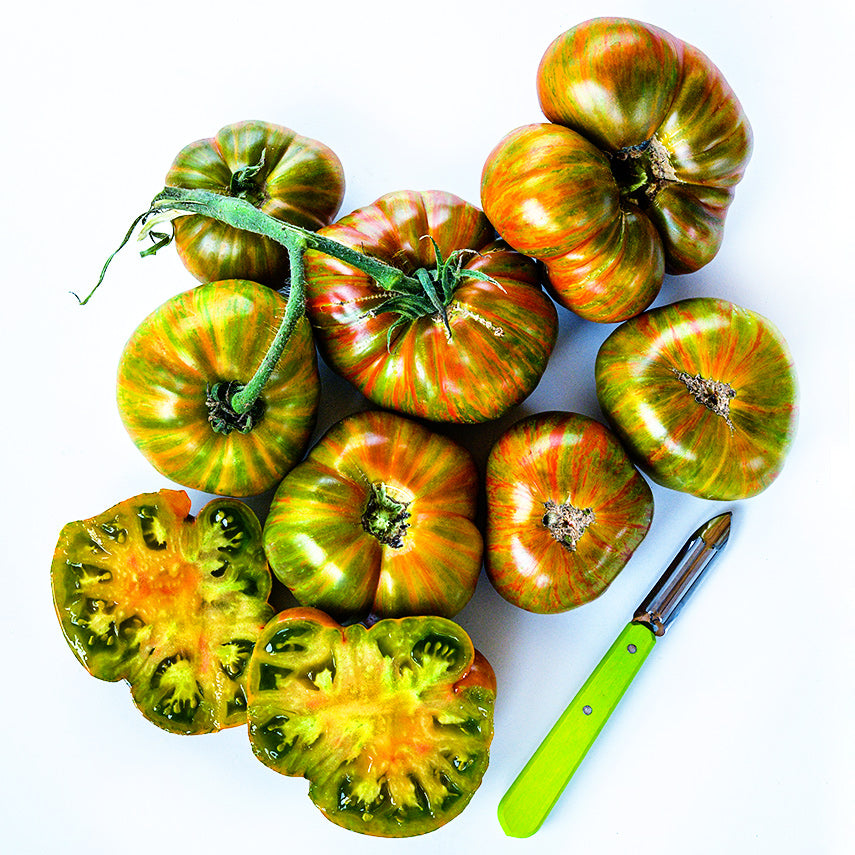 Seven whole Berkeley Tie-Dye Green Tomatoes arranged with a sliced tomato and green handled peeler.