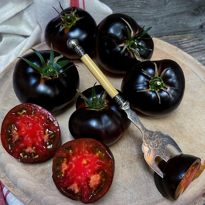 A half dozen whole and cut Black Beauty Tomatoes with a serving fork on a wood platter. Image by Lucy Hutchins.